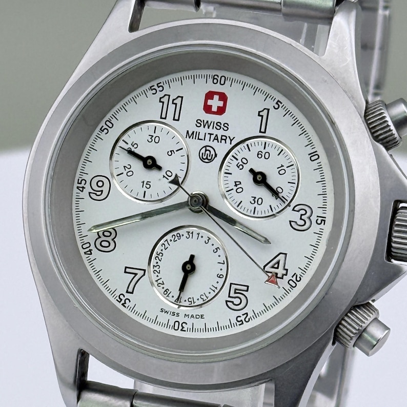 Puede incluir: Un reloj Swiss Military plateado con esfera blanca. El reloj presenta n&uacute;meros negros, tres subesferas y un logotipo de cruz suiza roja. Tiene una correa de acero inoxidable y funci&oacute;n cron&oacute;grafo.