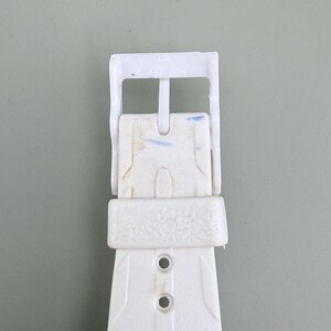 Puede incluir: Primer plano de una hebilla de correa de reloj blanca. La hebilla de pl&aacute;stico tiene forma rectangular con orificios de ajuste. La correa presenta un dise&ntilde;o texturizado y el texto "MOD DEP" es visible.