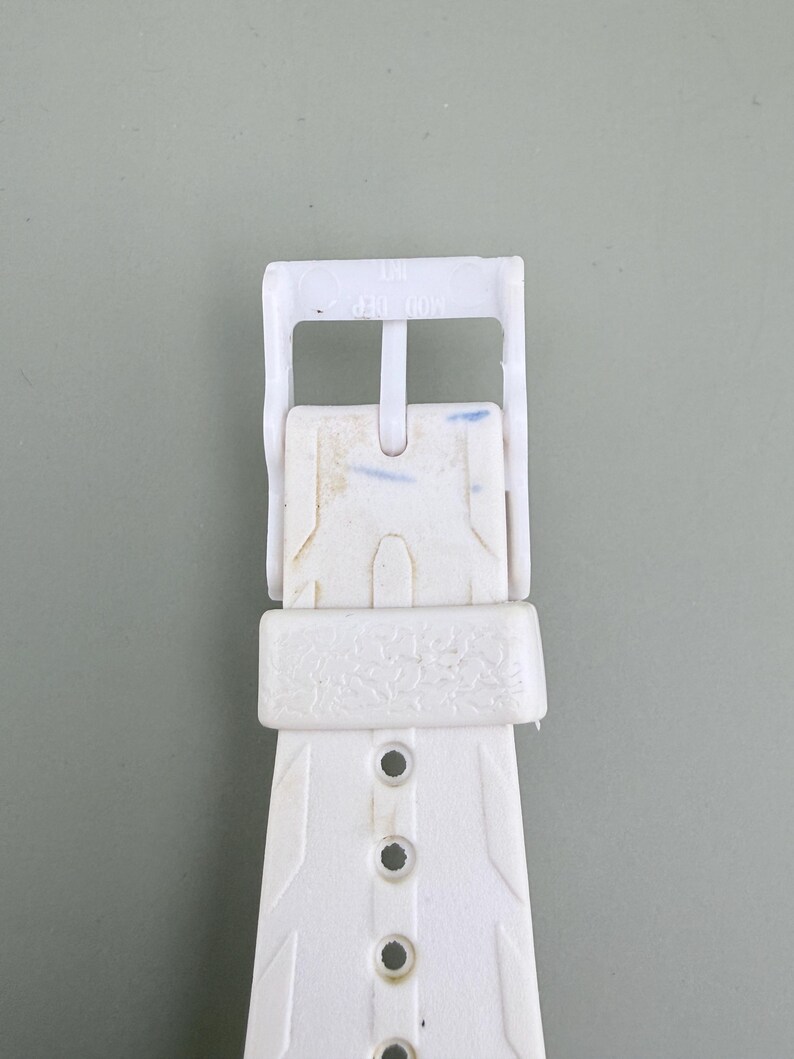 Puede incluir: Primer plano de una hebilla de correa de reloj blanca. La hebilla de pl&aacute;stico tiene forma rectangular con orificios de ajuste. La correa presenta un dise&ntilde;o texturizado y el texto "MOD DEP" es visible.
