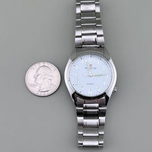 Pu&ograve; includere: Un orologio da polso argentato con quadrante bianco e cinturino argentato. Il quadrante mostra il marchio "BINCCHI" e la parola "QUARTZ". Una moneta &egrave; accanto all'orologio per il confronto delle dimensioni.