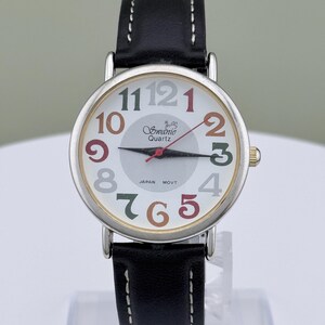 May include: A silver-toned quartz watch with a white face and a black leather band. The watch face features colorful numerals and the brand name "Swanic Quartz". The watch has a second hand and the text "JAPAN MOVT" is visible.