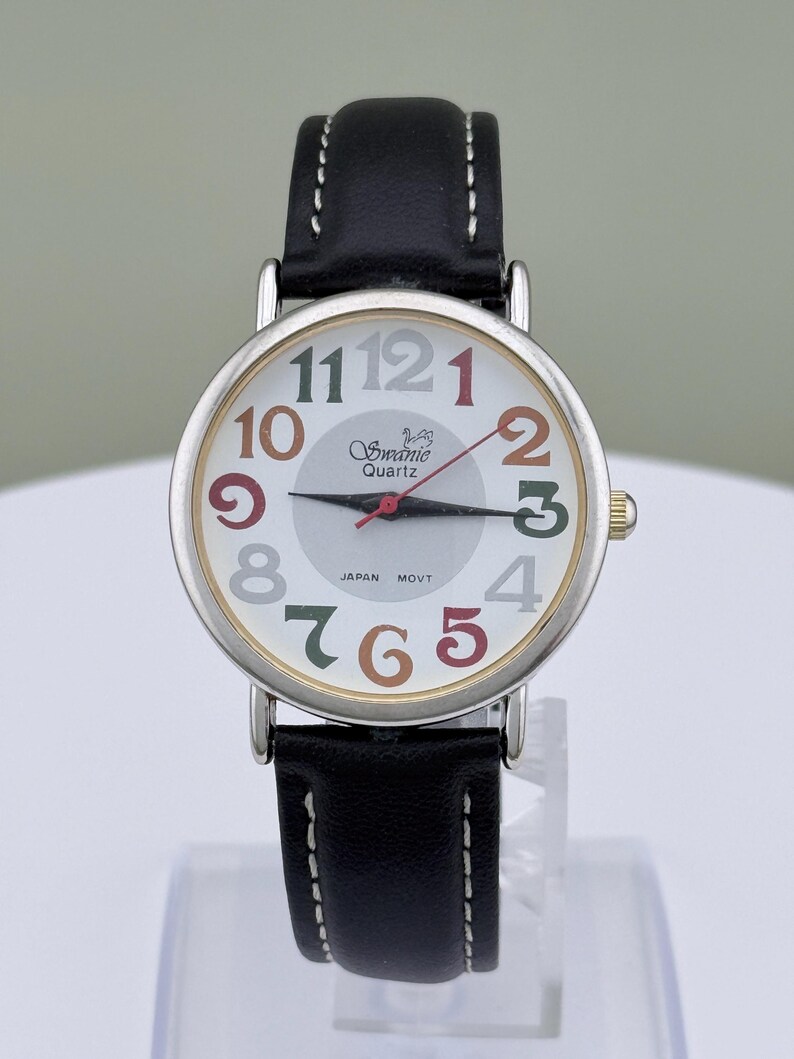 May include: A silver-toned quartz watch with a white face and a black leather band. The watch face features colorful numerals and the brand name "Swanic Quartz". The watch has a second hand and the text "JAPAN MOVT" is visible.