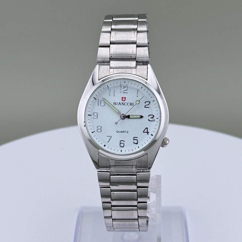 Pu&ograve; includere: Un orologio da polso argentato con quadrante bianco e cinturino in metallo abbinato. Il quadrante mostra il marchio "Biancchi" e la parola "Quartz". L'orologio ha un quadrante rotondo con numeri e lancette nere.