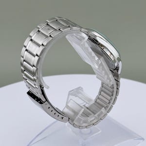 Pu&ograve; includere: Un orologio da polso argentato con cinturino in acciaio inossidabile. Il quadrante dell'orologio &egrave; visibile e il cinturino &egrave; collegato con una chiusura. L'orologio &egrave; esposto su un supporto di plastica trasparente.
