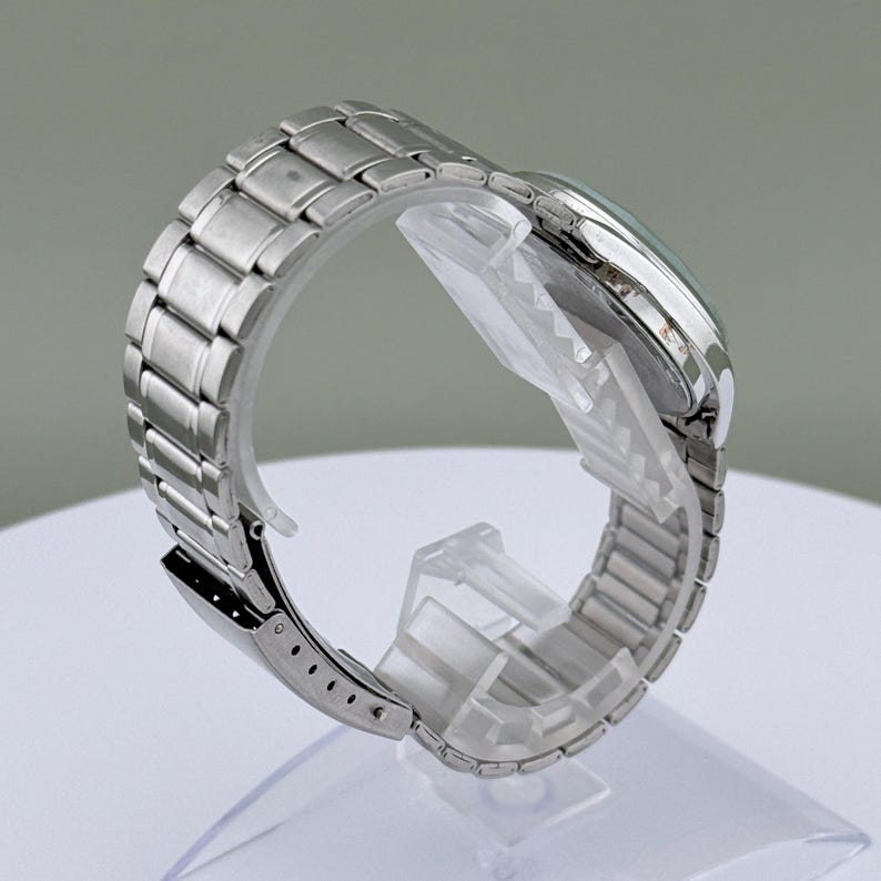 Pu&ograve; includere: Un orologio da polso argentato con cinturino in acciaio inossidabile. Il quadrante dell'orologio &egrave; visibile e il cinturino &egrave; collegato con una chiusura. L'orologio &egrave; esposto su un supporto di plastica trasparente.