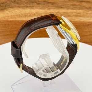 May include: A vintage wristwatch with a brown leather band and a gold-toned bezel. The watch face is rectangular with a silver and gold color scheme. The band is secured with a gold-colored buckle. The watch is displayed on a clear plastic stand.