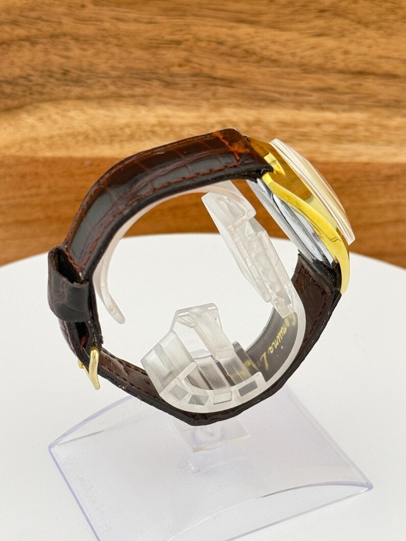 May include: A vintage wristwatch with a brown leather band and a gold-toned bezel. The watch face is rectangular with a silver and gold color scheme. The band is secured with a gold-colored buckle. The watch is displayed on a clear plastic stand.