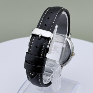 May include: A black leather watch band with white stitching and a silver buckle. The watch face is visible, with a silver case and a dark face. The watch is displayed on a clear stand.