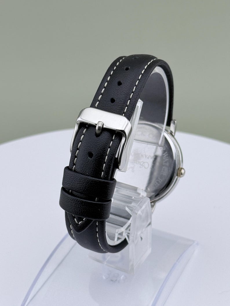 May include: A black leather watch band with white stitching and a silver buckle. The watch face is visible, with a silver case and a dark face. The watch is displayed on a clear stand.