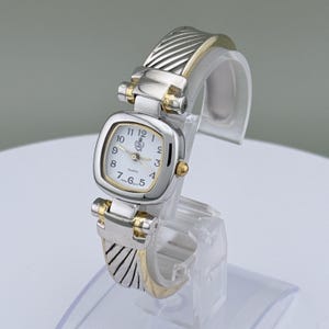 May include: A silver and gold-toned quartz watch with a square face. The watch features a white dial with black numerals and gold-toned hands. The band has a textured design. The watch is displayed on a clear stand.