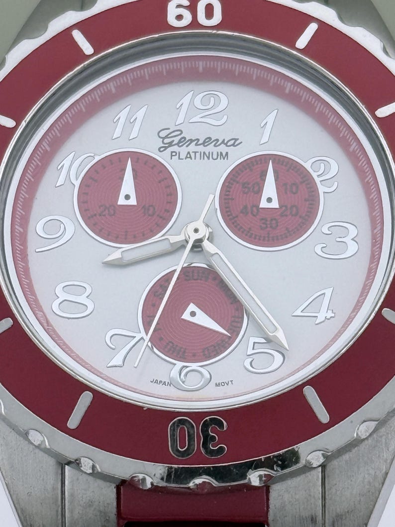 May include: A Geneva Platinum wristwatch with a silver face and red accents. The watch features three sub-dials, silver hands, and a red bezel. The numbers are white, and the watch has a stainless steel band.