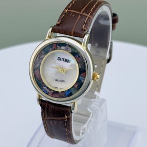 May include: A vintage Gitano quartz watch with a brown leather band. The watch face features a mother-of-pearl center surrounded by colorful stones and gold-tone accents. The watch is displayed on a clear stand.