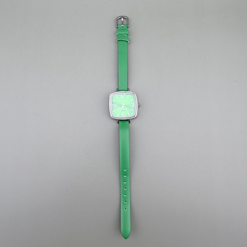 May include: A green wristwatch with a square face and a silver bezel. The watch has a light green face with white numerals and hands. The watchband is green leather. The watch is displayed against a grey background.