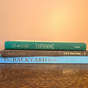 May include: A stack of three books with titles visible: "Yardening," "A Southern Album," and "The Backyard Book." The books are in shades of green, brown, and blue, stacked on a wooden surface. The top book is green with the title "Yardening."