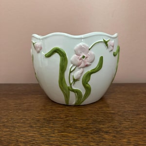 May include: A white ceramic planter with a scalloped rim, featuring raised, three-dimensional green stems and light pink floral accents embossed on the exterior surface. The vessel has a smooth, glossy finish and a rounded, bowl-like shape.
