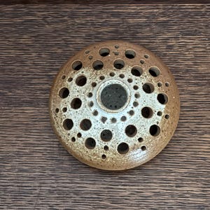 May include: A circular ceramic object with a speckled, brown and cream glaze. It features numerous circular holes of varying sizes, arranged in concentric circles. The central hole is larger. Likely a decorative piece or diffuser.