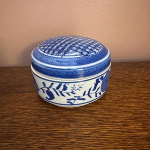May include: A round ceramic lidded jar featuring a white base with blue hand-painted floral and vine motifs. The lid displays a blue grid pattern on top, bordered by a geometric Greek key design, while the base shows blue botanical illustrations.