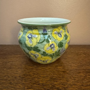 May include: A round ceramic planter featuring a hand-painted pattern of yellow pansies with purple centers and green leaves against a light green wash background. The pot has a wide, flared rim and a smooth, glossy finish throughout the exterior.