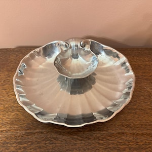 Puede incluir: Una fuente para servir plateada en forma de concha con un plato central más pequeño. El plato tiene un borde festoneado y una superficie reflectante, adecuado para servir aperitivos o salsas. El plato está colocado sobre una superficie de madera.