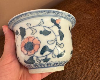 May include: A white ceramic bowl with a blue floral design and a pink flower. The bowl has a blue rim and is held in a hand. The bowl is likely for decorative purposes.