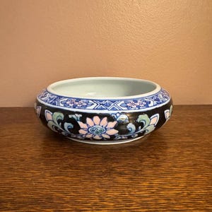 Puede incluir: Un cuenco de cerámica pequeño y redondo con un exterior negro adornado con patrones florales y geométricos en azul, rosa y verde. El interior es blanco y el borde presenta un diseño azul y blanco. El cuenco está sobre una superficie de madera.