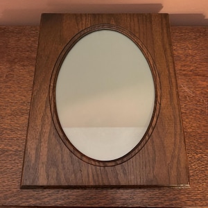 May include: A rectangular wooden frame with a dark brown finish and a prominent wood grain pattern. The frame features a central oval-shaped opening that contains a smooth, reflective mirror surface, creating a contrast between the wood and glass.
