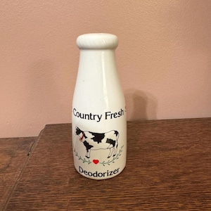May include: White ceramic deodorizer in the shape of a milk bottle. The bottle features a black and white cow illustration with the words "Country Fresh" and "Deodorizer". A red heart is below the cow.