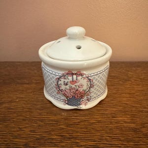 May include: A white ceramic jar with a lid featuring a floral basket design. The jar has a round shape with a decorative pattern in blue and gray. The floral design includes pink roses and a red bow.