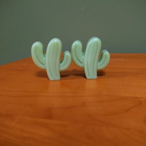Puede incluir: Dos objetos decorativos en forma de cactus verde claro. Los cactus están hechos de un material translúcido y se asientan sobre una superficie de madera. El fondo es verde liso y apagado. Estos cactus son un artículo de decoración único.