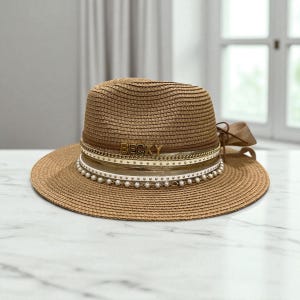 Puede incluir: Sombrero fedora de paja marrón con ala ancha. El sombrero presenta una cinta marrón, una cadena dorada, una banda blanca con perlas y el nombre "BECKY" en letras doradas. Un lazo de cinta marrón está atado a un lado.