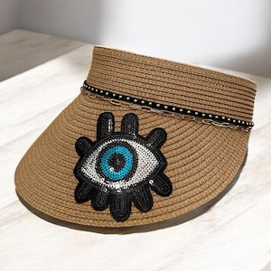 Può includere: Visiera in paglia marrone con un motivo a occhio diabolico decorativo. La visiera ha un dettaglio a catena nera e argento. L'occhio diabolico è nero con un centro di paillettes blu e argento.