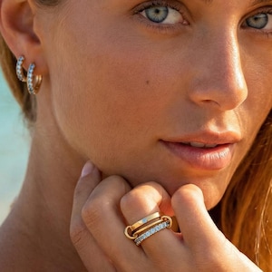 Può includere: Una donna indossa orecchini a cerchio dorati e un anello d'oro con una fascia di diamanti. L'anello è sul dito della donna. La donna ha i capelli biondi e gli occhi azzurri. Lo sfondo è una spiaggia con acqua blu.