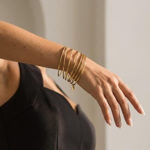 以下が含まれることがあります： 手首に金色のバングルがいくつかあります。バングルは細くて円形で、小さな長方形のチャームが1つにぶら下がっています。その人は黒いトップスを着ています。