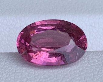 May include: A faceted, oval-shaped gemstone in shades of pink and magenta. The gem has a polished surface with internal inclusions, set against a neutral background. The gem is approximately 2.5 cm in length.