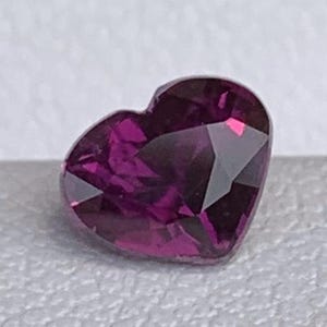 Puede incluir: Una gema facetada en forma de corazón, de un intenso color magenta. La gema se presenta sobre un fondo neutro, resaltando sus superficies pulidas y sus reflejos internos. El corte de la piedra destaca su forma y color.