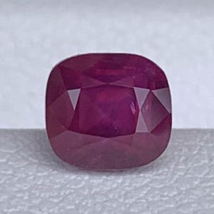 May include: A cushion-cut ruby gemstone, displaying a deep red hue. The gemstone is faceted, with a square shape and rounded corners. The ruby is set against a white background, highlighting its color and clarity.