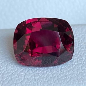 May include: A cushion-cut, deep red gemstone with facets reflecting light. The gem is set against a white, textured background, highlighting its color and clarity. This gemstone is likely for jewelry.