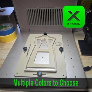 May include: A silver-colored metal device with a beige-colored insert, featuring the text "AngelTechLab.com" and "F1/F2 U PMAG 30 M2/M3". The image also includes a green square with the white text "XTOOL" and the phrase "Multiple Colors to Choose".