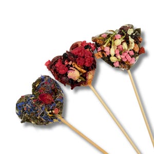 May include: Three heart-shaped tea arrangements on wooden sticks. The first features blue cornflowers and red rose petals. The second contains red berries and rose buds. The third is topped with white jasmine flowers and small pink fruit cubes.