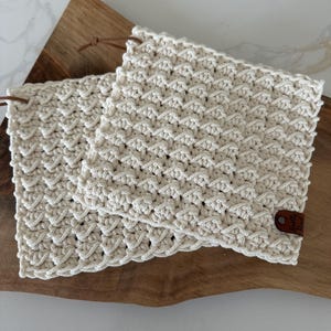 May include: Two cream-coloured, textured crochet potholders with a leather tag and loop. They are placed on a wooden chopping board. The crochet pattern creates a raised, geometric design.