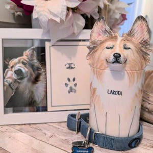 Op de afbeelding: Een keramische dierenurne in de vorm van een hond met bruin en wit haar. De urne heeft de naam "Lakota" erop geschreven. Een blauwe hondenriem met een label met de tekst "Lakota" ligt naast de urne. Op de achtergrond is een ingelijste foto van een hond te zien.
