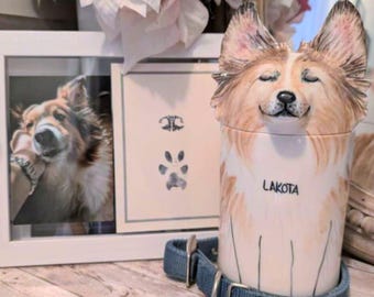May include: A ceramic pet urn shaped like a dog with brown and white fur. The urn has the name "Lakota" written on it. A blue dog collar with a tag that says "Lakota" is next to the urn. A framed photo of a dog is in the background.