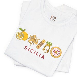 May include: A white t-shirt featuring a graphic print with a yellow lemon, a Trinacria symbol, a cannolo, a crowned figure, and a red cart wheel. Below the icons, the word SICILIA is printed in red capital letters. The shirt has a round neckline.