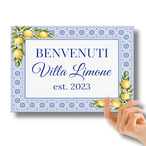 May include: Rectangular sign with the text "BENVENUTI Villa Limone est. 2023" in blue script. The sign has a white background with a blue floral border and lemon illustrations. The sign is being held by a hand.