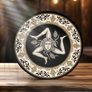 May include: A round decorative plate featuring the Trinacria symbol, a figure with three legs and a face, in the center. The design is black, white, and gold, with an ornate border. The plate is set on a wooden surface.