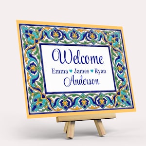 K&ouml;nnte beinhalten: Dekoratives Schild mit gelbem Rand und blau-gr&uuml;nem Blumenmuster. Auf dem Schild steht "Willkommen" in blauer Schrift, gefolgt von Namen und dem Nachnamen "Anderson". Das Schild wird auf einer kleinen Holzstaffelei pr&auml;sentiert.