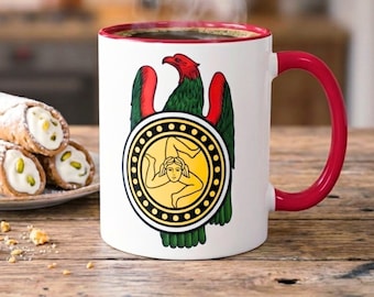May include: A white ceramic mug with a red interior and handle, filled with coffee and steam. The mug features a colorful emblem with a red eagle, green wings, and a yellow circular design. Cannoli pastries are on a plate.