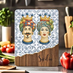 May include: A wooden chopping board with a blue and white patterned tile design featuring two stylised faces with crowns. The text "Sicilia" is written on the wood.