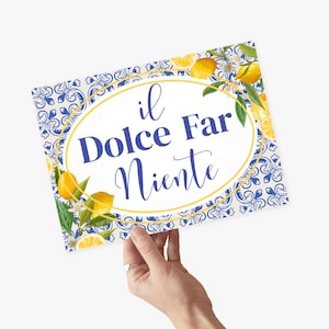 Puede incluir: Un cartel blanco con un borde estampado en azul y blanco y limones. El cartel tiene un óvalo dorado con el texto "il Dolce Far Niente" en cursiva azul.