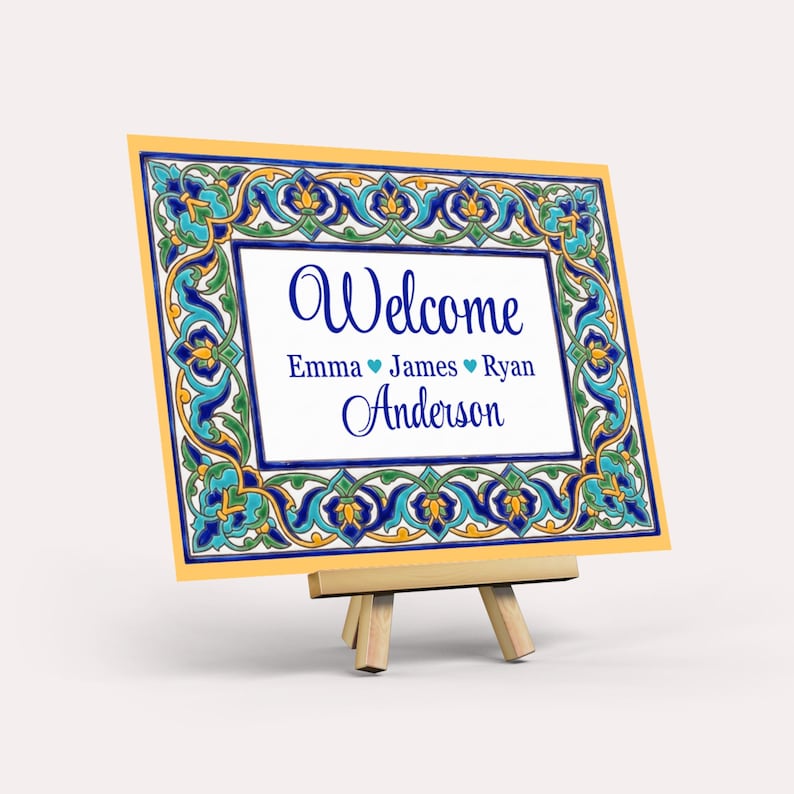 K&ouml;nnte beinhalten: Dekoratives Schild mit gelbem Rand und blau-gr&uuml;nem Blumenmuster. Auf dem Schild steht "Willkommen" in blauer Schrift, gefolgt von Namen und dem Nachnamen "Anderson". Das Schild wird auf einer kleinen Holzstaffelei pr&auml;sentiert.